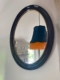 Le Miroir ovale noir 1950 reflète un abat-jour sombre avec une frange orange et fait partie d'une pièce lumineuse et confortable, évoquant l'élégance intemporelle des années 1950. Brocante 93