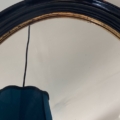 Un gros plan du Miroir ovale noir 1950, doté d'un cadre noir et or, reflète une partie d'un abat-jour et d'un plafond bleus. Cette pièce élégante évoque le style intemporel des années 1950. Brocante 93