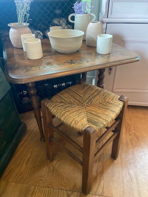 Petite table en bois avec tasses et bols en céramique, à côté d'un charmant Tabouret paille vintage 1960 avec siège tressé, posé sur un sol en bois. Brocante 93