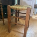 Un Tabouret paille vintage 1960 avec une assise tressée est posé sur un sol en bois à côté d'une table et d'un meuble. Brocante 93