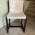 Une Chaise Gautier 1970, une chaise moderne blanche avec des pieds en bois foncé, se trouve près de l'escalier et d'un mur décoré d'œuvres d'art. Brocante 93