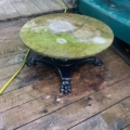 La Table basse début XXème siècle, une table ronde au plateau moussu et au piètement noir orné, est posée sur une terrasse en bois près d'un banc vert. Brocante 93