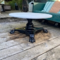 La Table basse début XXème siècle, une table ronde en marbre blanc avec un pied en griffe noir, est placée sur une terrasse en bois usé par le temps avec des sièges extérieurs en arrière-plan. Brocante 93