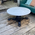 Table basse début XXème siècle : plateau rond en marbre avec base noire ornée, posé sur une terrasse en bois à côté d'un canapé vert avec des coussins rayés. Brocante 93