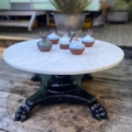 La Table basse début XXème siècle, ronde et blanche, est surmontée de petites plantes en pot et placée sur une terrasse en bois au milieu d'une végétation luxuriante. Brocante 93