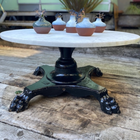 La Table basse début XXème siècle est une table ronde blanche avec un pied griffe noir, installée à l'extérieur sur une terrasse en bois et présentant cinq petites plantes en pot. Brocante 93