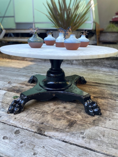 La Table basse début XXème siècle est une table ronde blanche avec un pied griffe noir, installée à l'extérieur sur une terrasse en bois et présentant cinq petites plantes en pot. Brocante 93