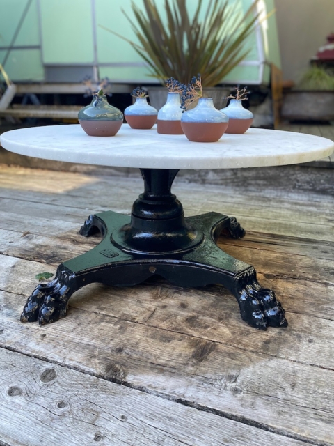 La Table basse début XXème siècle est une table ronde blanche avec un pied griffe noir, installée à l'extérieur sur une terrasse en bois et présentant cinq petites plantes en pot. Brocante 93