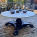 La Table basse début XXème siècle présente un plateau rond en marbre et une base noire ornée, évoquant le style du début du XXème siècle. Quatre petits vases décorent la surface. Elle est placée sur une terrasse extérieure en bois. Brocante 93