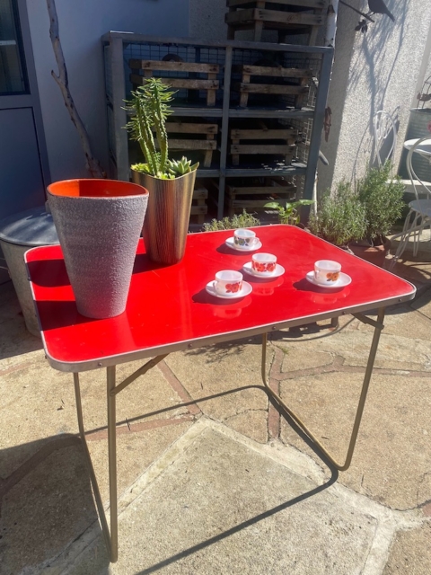 Une table pliante Lafuma 1970 en rouge est exposée au soleil, accompagnée d'une plante, d'un vase gris et de quatre petites tasses à thé sur des soucoupes. Brocante 93
