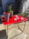 Une table pliante Lafuma 1970 en rouge est exposée au soleil, accompagnée d'une plante, d'un vase gris et de quatre petites tasses à thé sur des soucoupes. Brocante 93