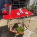 Une table pliante rouge Lafuma 1970 avec trois tasses à thé, des soucoupes et un grand vase se trouve à côté d'une jardinière de succulentes à l'extérieur. Brocante 93