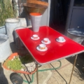 Une table pliante Lafuma 1970 en rouge est exposée au soleil, décorée de quatre tasses à thé, de soucoupes, de fraises et d'un grand vase gris. Brocante 93