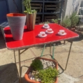 Une table pliante Lafuma 1970 accueille des tasses et des vases à l'extérieur, tandis que des plantes grasses en pot sont disposées sur l'étagère inférieure. Brocante 93