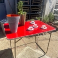 Une Table pliante Lafuma 1970 en rouge, placée à l'extérieur avec une plante, un vase gris et quatre petits services à thé sur le dessus, apporte un charme vintage et une touche des années 1970 - idéal pour recréer une ambiance authentique inspirée de Lafuma. Brocante 93