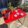 Une Table pliante Lafuma 1970 rouge vif présente un cactus en pot, un grand vase et quatre tasses à espresso avec soucoupes à l'extérieur. Brocante 93