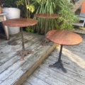 Trois tables rondes Guéridon de bistrot 1950 avec des bases en métal sont posées sur une terrasse en bois, entourées de plantes vertes luxuriantes. Brocante 93