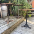 Trois tables en métal Guéridon de bistrot 1950 sont posées sur une terrasse en bois, entourées de plantes vertes et de décorations d'extérieur. Brocante 93