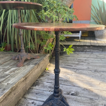 Le Guéridon de bistrot 1950 est une table ronde avec un piètement en métal orné, posée sur une terrasse en bois, entourée de plantes vertes et de murs colorés. Brocante 93