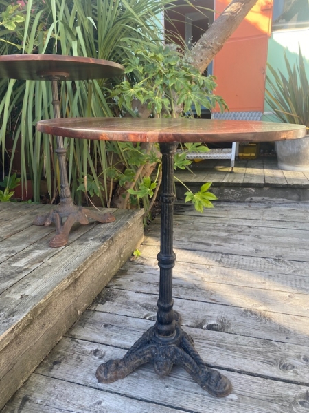 Le Guéridon de bistrot 1950 est une table ronde avec un piètement en métal orné, posée sur une terrasse en bois, entourée de plantes vertes et de murs colorés. Brocante 93