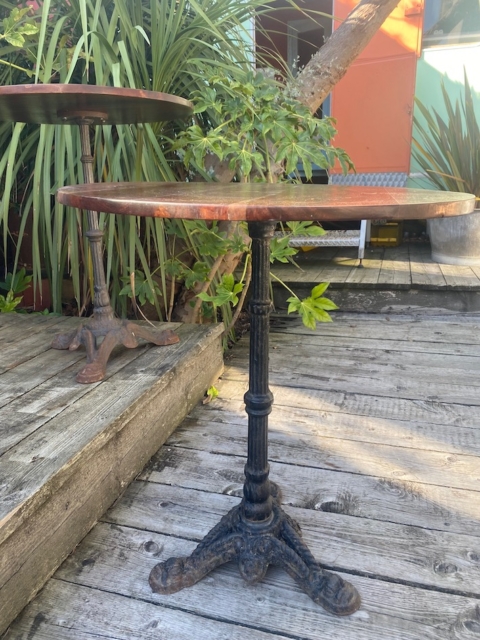 Le Guéridon de bistrot 1950 est une table ronde avec un piètement en métal orné, posée sur une terrasse en bois, entourée de plantes vertes et de murs colorés. Brocante 93