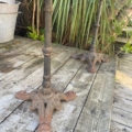 Deux pieds de table en métal rouillé et orné, provenant d'un Guéridon de bistrot 1950, reposent sur une terrasse en bois usée par les intempéries, avec une verdure luxuriante en arrière-plan. Brocante 93