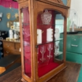 La Vitrine murale vintage 1950 présente des étagères en verre qui exposent élégamment des verres, des bougies et un bol en cristal sur un fond rouge audacieux. Brocante 93