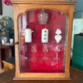 Vitrine murale vintage 1950 avec portes en verre, exposant des bougies, de la verrerie et un pot décoratif sur un fond rouge saisissant. Brocante 93