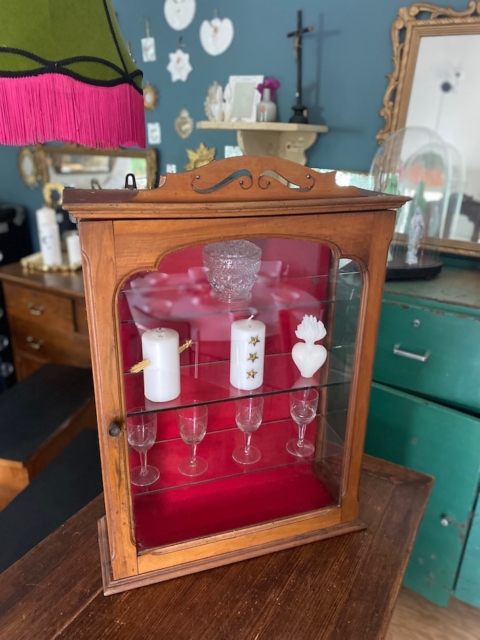Une Vitrine murale vintage 1950 présente des bougies et de la verrerie, élégamment disposées sur une table dans une charmante pièce de style rétro. Brocante 93