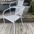 La Paire de fauteuils de jardin blancs en métal est disposée sur une terrasse en bois, entourée de plantes et d'objets de jardin en arrière-plan. Brocante 93