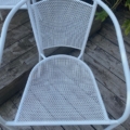 Une paire de fauteuils de jardin en métal blanc est posée sur une terrasse en bois usée par le temps, encadrée par des plantes vertes luxuriantes en arrière-plan. Brocante 93
