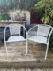 La paire de fauteuils de jardin blancs en métal, placée sur une terrasse en bois et entourée de plantes, offre un espace convivial et idéal pour profiter du plein air. Brocante 93