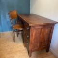 Une chaise en bois est assise à côté du Bureau vintage 1960, un bureau en bois avec trois tiroirs, dans le coin d'une pièce. Brocante 93