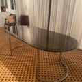 La Table verre & chrome 1970, dotée d'un plateau en verre et de pieds chromés élégants, est posée sur un tapis à motifs dans une pièce aux rideaux blancs. Brocante 93