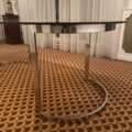 La Table verre & chrome 1970 présente un plateau rond en verre et une élégante base chromée, placés sur un tapis à motifs bruns et des rideaux blancs, dans un style chic des années 1970. Brocante 93