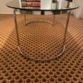 La Table verre & chrome 1970, dotée de pieds chromés, est posée sur un tapis marron à motifs au milieu d'étagères, évoquant un style rétro chic. Brocante 93