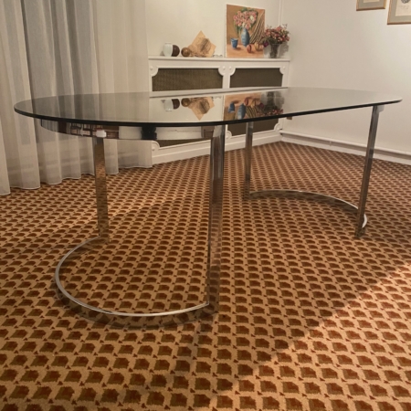 La Table verre & chrome 1970, avec son plateau ovale en verre noir et ses pieds chromés, se pose sur un tapis à motifs dans un salon à la décoration chaleureuse, créant un style rétro chic. Brocante 93