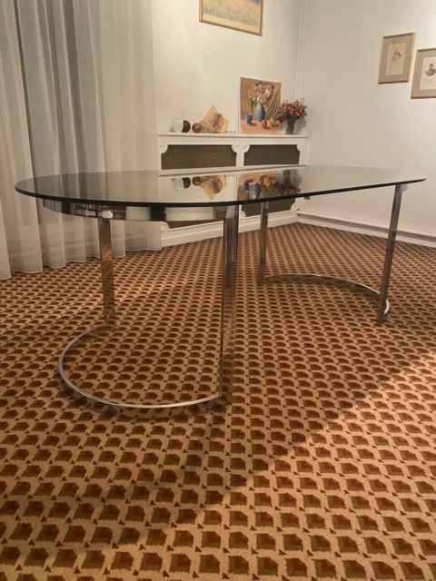 La Table verre & chrome 1970, avec son plateau ovale en verre noir et ses pieds chromés, se pose sur un tapis à motifs dans un salon à la décoration chaleureuse, créant un style rétro chic. Brocante 93