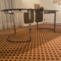 La Table verre & chrome 1970 est posée sur un tapis à motifs dans une pièce décorée d'œuvres d'art encadrées et de rideaux transparents. Brocante 93