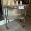La Desserte médicale vintage 1970, un chariot roulant en métal surmonté de quatre bocaux blancs, est placée à l'intérieur, près d'un escalier et d'un mur peint. Brocante 93