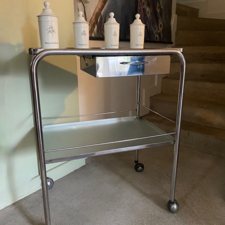 La Desserte médicale vintage 1970, avec quatre bocaux sur le dessus, se trouve sur un sol en béton près d'un escalier et d'une peinture murale. Brocante 93