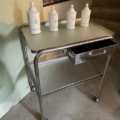 Desserte médicale vintage 1970, chariot à roulettes en métal avec quatre bocaux en céramique sur le dessus et un tiroir ouvert en dessous, présenté sur un sol en béton. Brocante 93