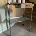 La Desserte médicale vintage 1970 est un chariot métallique sur quatre roues, contenant trois bocaux blancs, et présenté près d'un escalier contre un mur vert pâle. Brocante 93