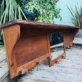 La Porte-manteaux bois 1940, une étagère murale en bois vintage avec des crochets, est exposée sur une terrasse en bois au milieu de plantes vertes luxuriantes. Brocante 93
