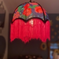 La Baladeuse bohème chic rose est un abat-jour floral coloré avec une frange rouge qui brille chaleureusement dans une pièce peu éclairée. Brocante 93