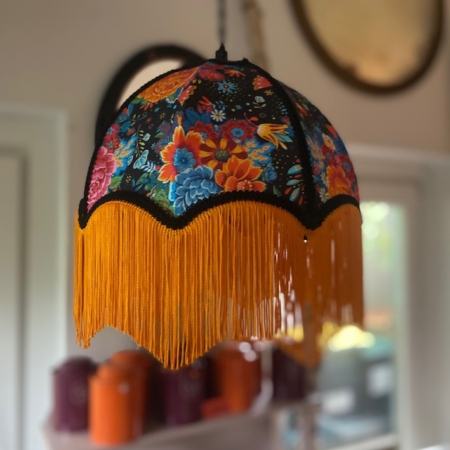 La Baladeuse bohème chic rose, un abat-jour floral avec une frange jaune, est suspendue à l'intérieur avec des étagères et un décor flous en arrière-plan. Brocante 93