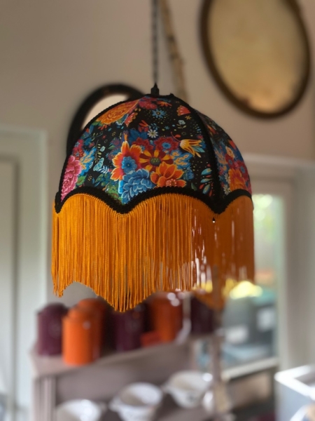 La Baladeuse bohème chic rose, un abat-jour floral avec une frange jaune, est suspendue à l'intérieur avec des étagères et un décor flous en arrière-plan. Brocante 93