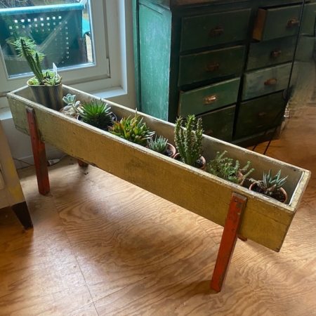 La jardinière Jardinières en bois 1950, remplie de plantes grasses, repose sur des pieds rouges audacieux près d'une fenêtre et d'une commode verte à l'intérieur, apportant une touche de style des années 1950. Brocante 93