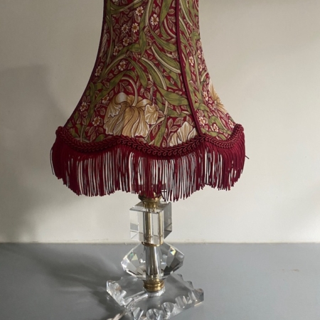 La Lampe pied en verre vintage 1940 présente un abat-jour à motifs floraux avec des franges rouges et un socle en verre clair à facettes, idéal pour ajouter un charme intemporel à n'importe quelle pièce. Brocante 93