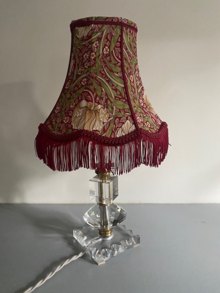 La Lampe pied en verre vintage 1940 présente un abat-jour à motifs floraux avec des franges rouges et un socle en verre clair à facettes, idéal pour ajouter un charme intemporel à n'importe quelle pièce. Brocante 93